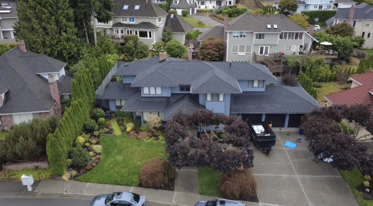 Aerial drone view of a multi-gabled suburban home with a newly installed dark gray shingle roof, construction trailer, and landscaped yard in Bellevue, Washington