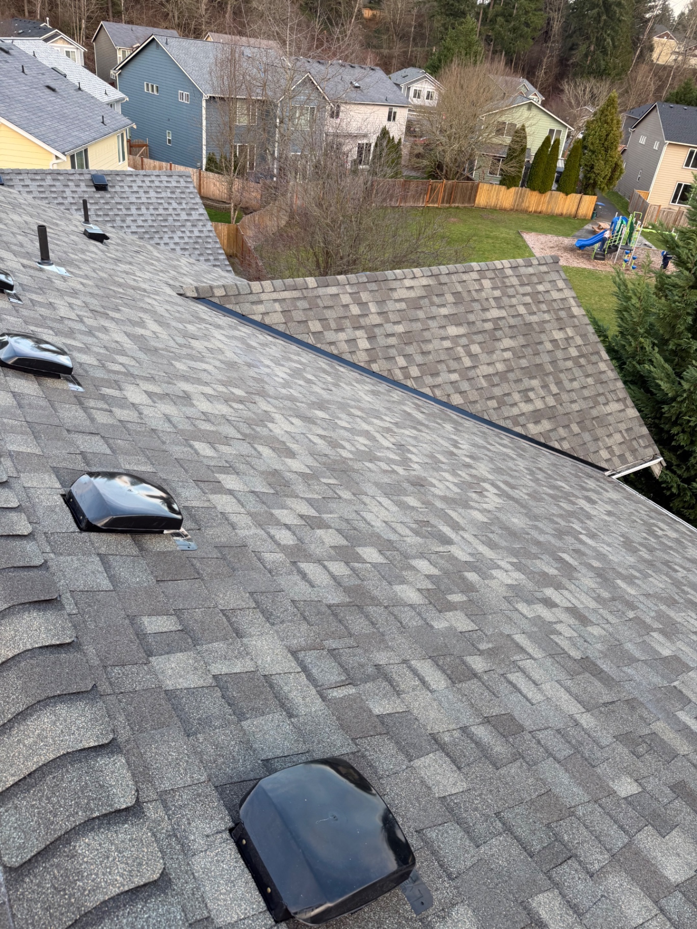 Angled view of a residential roof with multi-tonal gray architectural shingles, black vents, and flashing valleys overlooking a suburban neighborhood