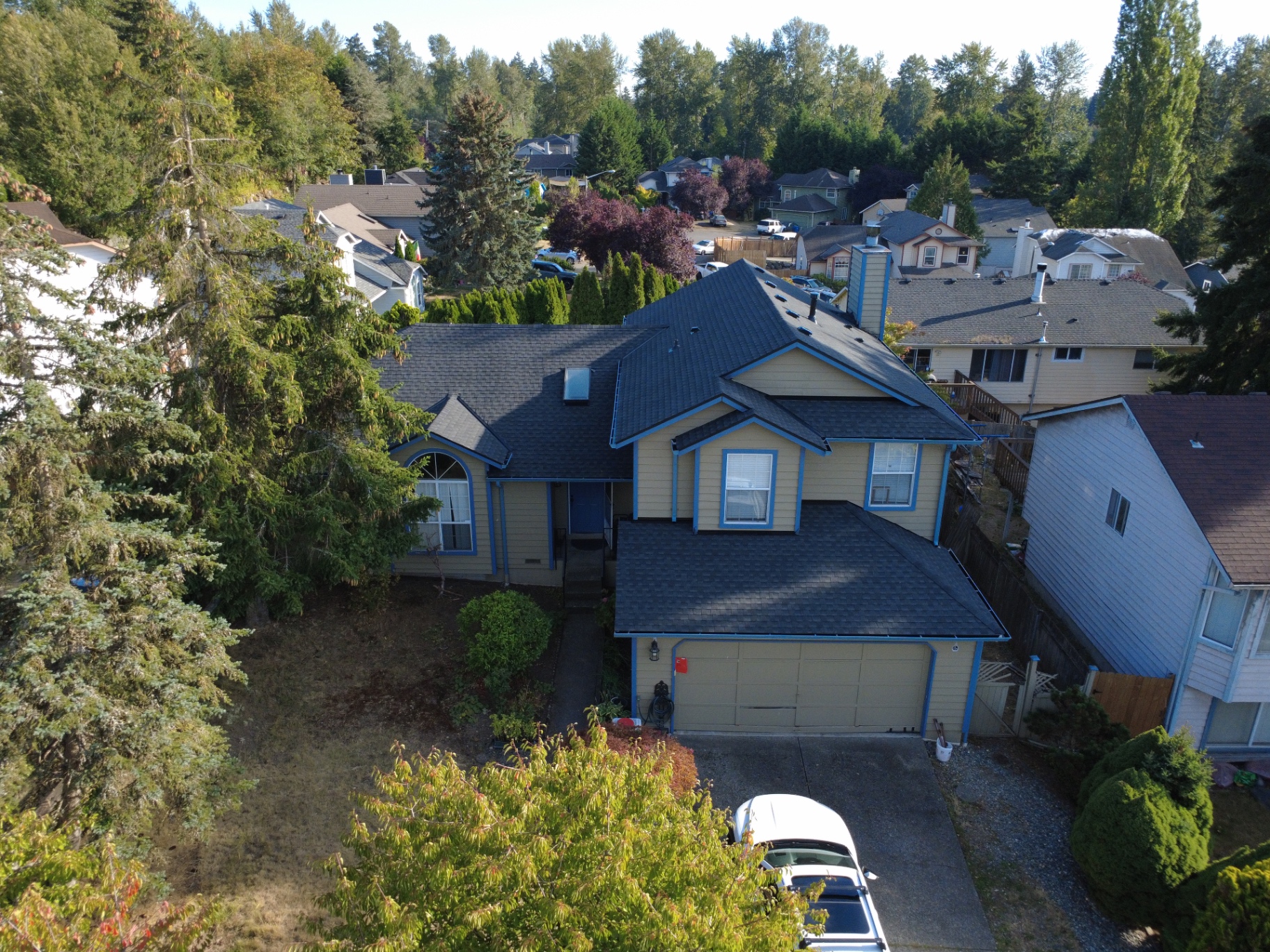 Front-facing high-angle drone view of a tan and blue-trimmed house with a fresh dark grey roof and matching gutters framed by lush green trees in Kent, Washington