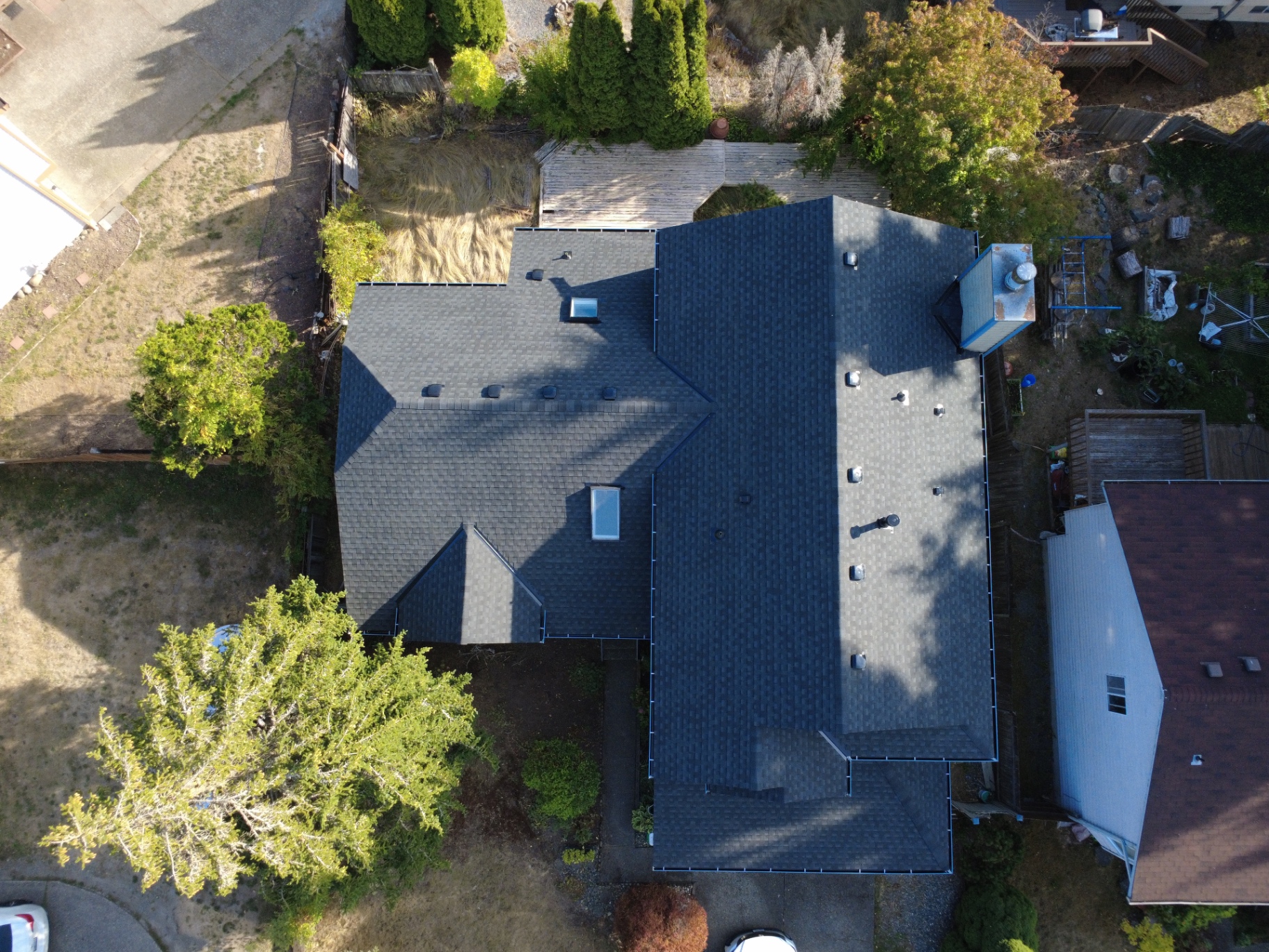 Overhead drone plan view of a complex dark grey shingled roofline with blue gutters, two skylights, and a light wood deck below at a Kent, Washington home