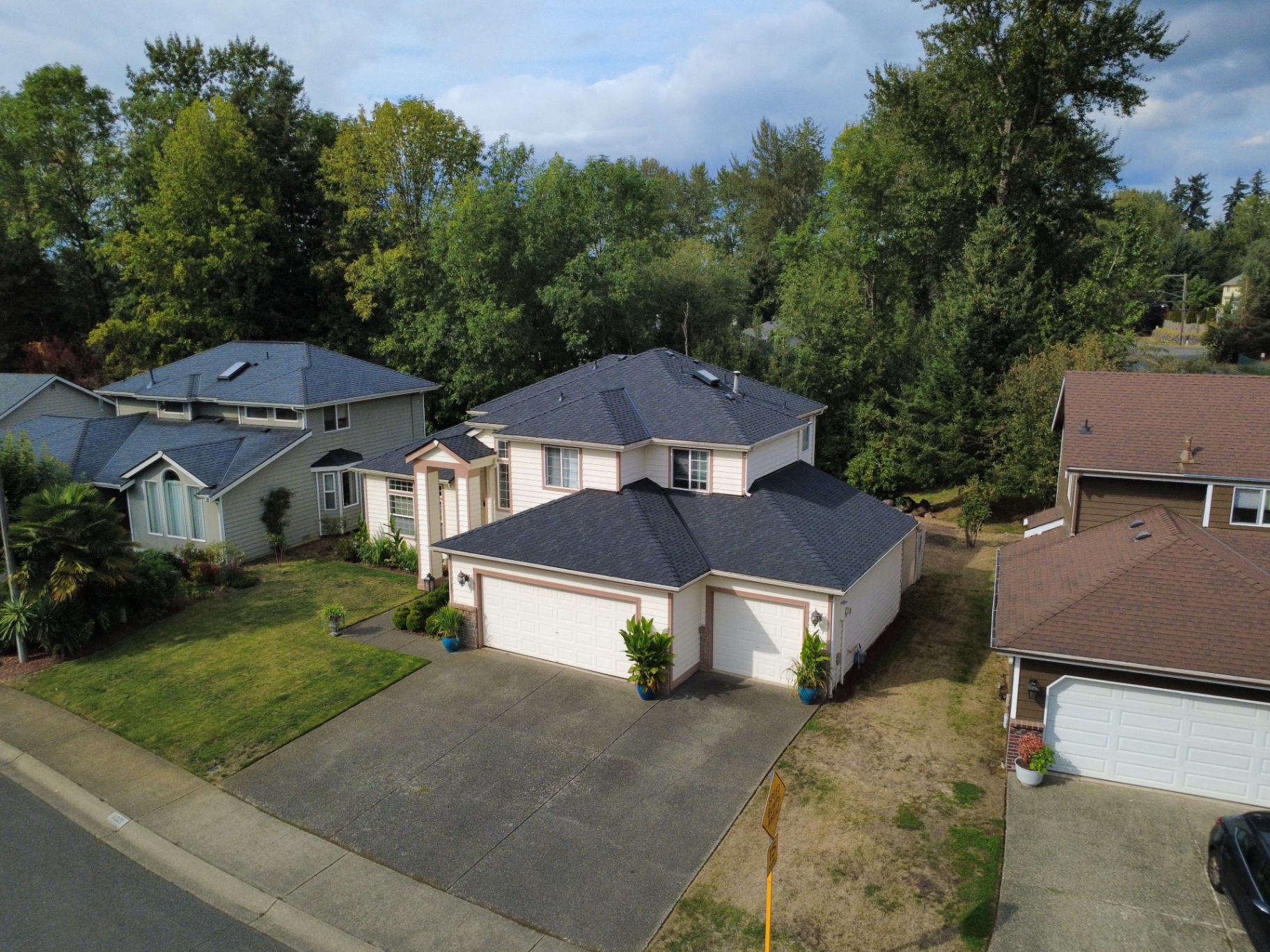 Oblique aerial drone view of the front and side of a cream-sided home with a new dark charcoal shingle roof and neighboring houses in Covington, Washington