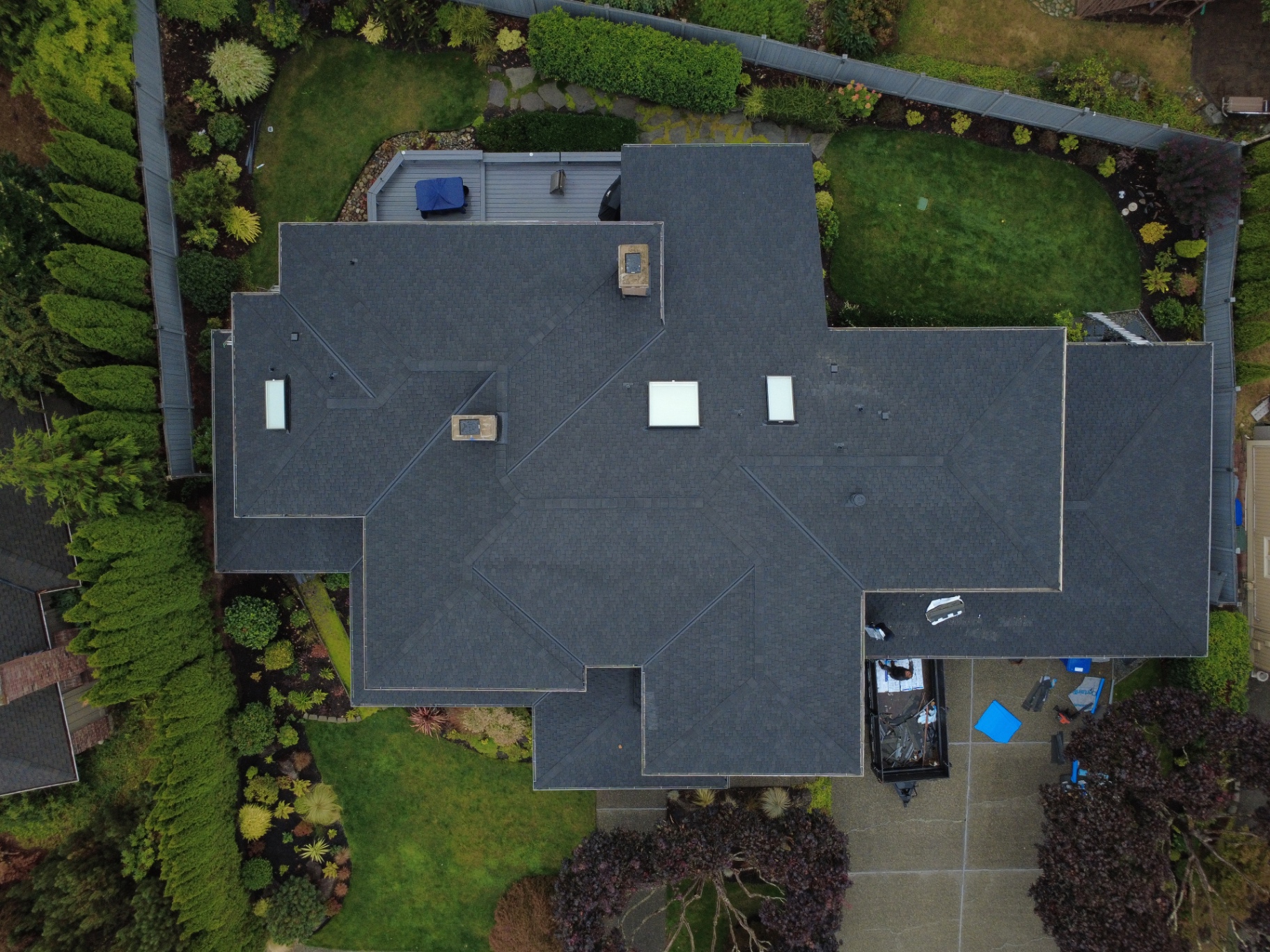 Top-down drone view of a complex dark gray shingle roof with skylights and chimneys, plus a rear wooden deck in Bellevue, Washington